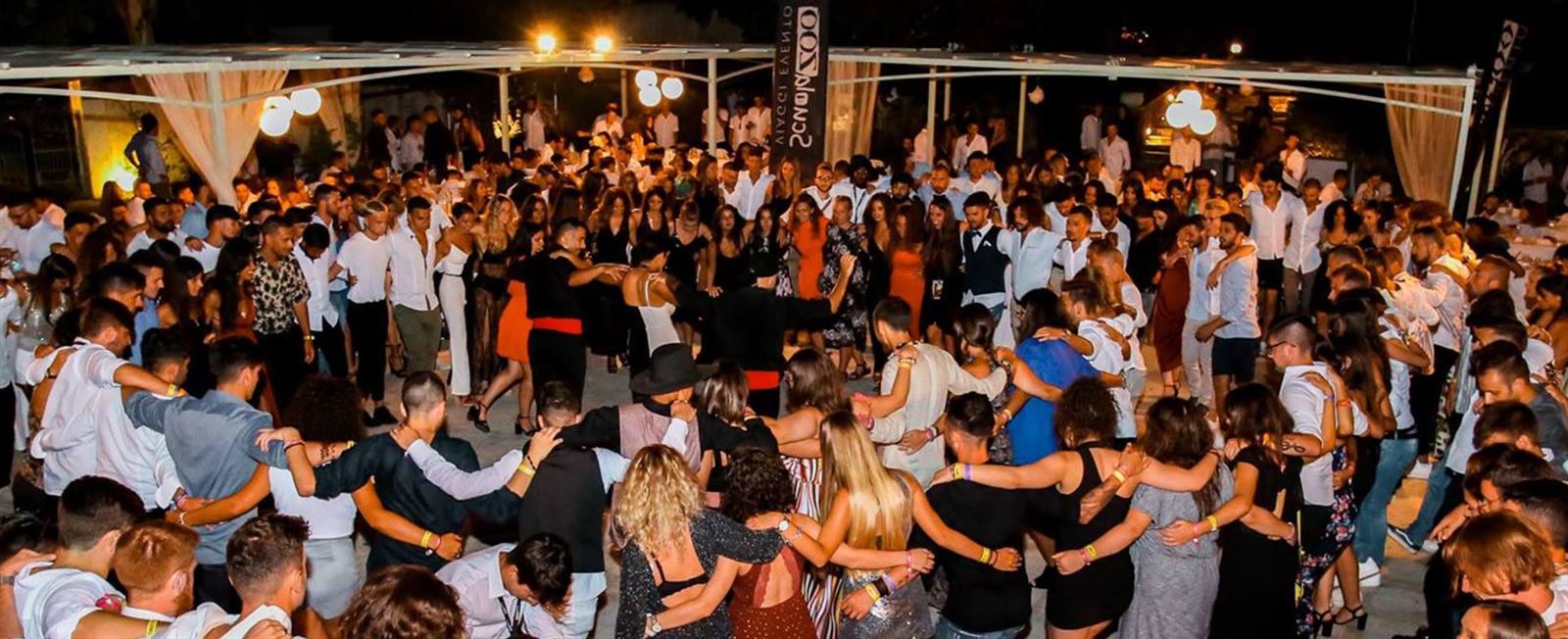 A large group dancing in a circle at an evening gathering.